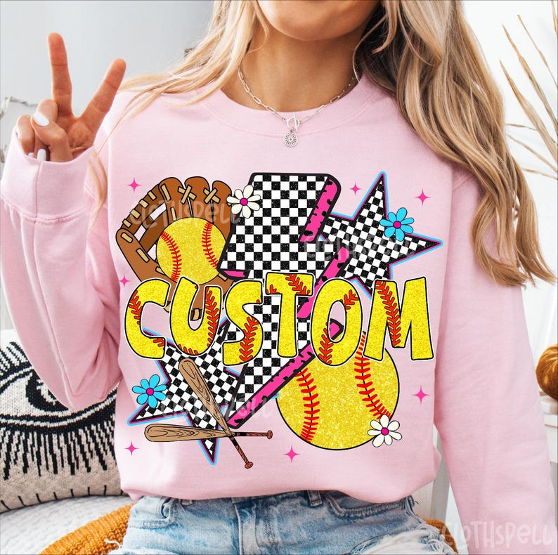 May include: Pink sweatshirt with a softball-themed graphic. The design includes a yellow softball, a brown glove, crossed bats, and the word "CUSTOM" in yellow. Accents include a checkered star, pink lightning bolt, and blue and white flowers.