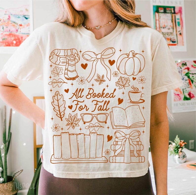 May include: Cream-colored t-shirt featuring a brown graphic design with fall-themed illustrations. The design includes a scarf, bow, pumpkin, leaves, glasses, coffee cup, open book, and stacked books. Text on the shirt reads "All Booked For Fall."
