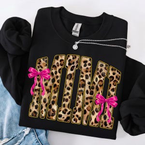 May include: Black sweatshirt with the word "MIMI" in leopard print letters outlined in gold. Pink bows are attached to the letters. A silver necklace with a pendant is draped across the sweatshirt.