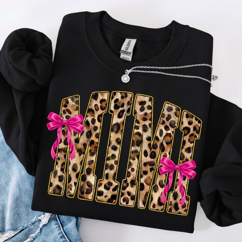 May include: Black sweatshirt with the word "MIMI" in leopard print letters outlined in gold. Pink bows are attached to the letters. A silver necklace with a pendant is draped across the sweatshirt.