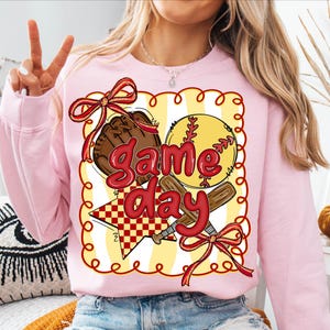 Puede incluir: Una sudadera rosa claro con un dise&ntilde;o tem&aacute;tico de b&eacute;isbol. El dise&ntilde;o incluye una pelota de b&eacute;isbol, un guante y un bate, con las palabras "Game Day" en rojo. El dise&ntilde;o est&aacute; enmarcado con un borde amarillo y lazos rojos.