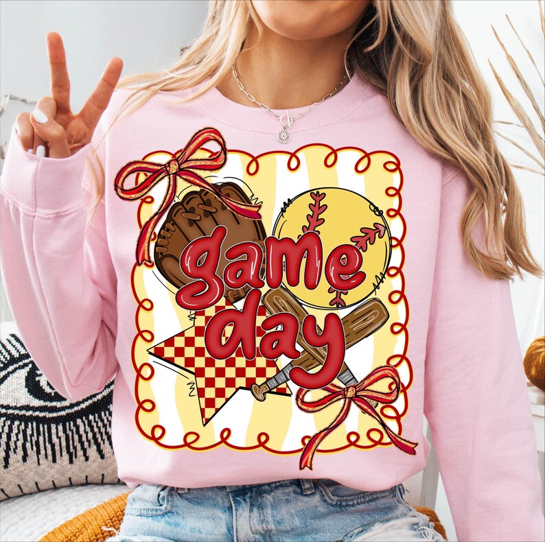 Puede incluir: Una sudadera rosa claro con un dise&ntilde;o tem&aacute;tico de b&eacute;isbol. El dise&ntilde;o incluye una pelota de b&eacute;isbol, un guante y un bate, con las palabras "Game Day" en rojo. El dise&ntilde;o est&aacute; enmarcado con un borde amarillo y lazos rojos.