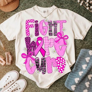 May include: Cream-colored t-shirt with the words "FIGHT HOPE CURE" in pink, decorated with a pink ribbon, bows, and daisy accents. The letters have various patterns, including polka dots, leopard print, and gingham.