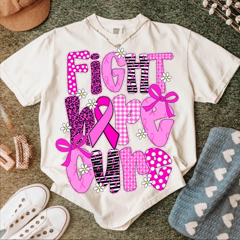 May include: Cream-colored t-shirt with the words "FIGHT HOPE CURE" in pink, decorated with a pink ribbon, bows, and daisy accents. The letters have various patterns, including polka dots, leopard print, and gingham.