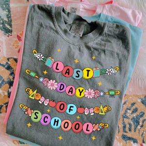 May include: A stack of three Comfort Colors t-shirts in gray, pink, and light blue. The gray shirt features the text "LAST DAY OF SCHOOL" in colorful bubble letters, with school-themed graphics.