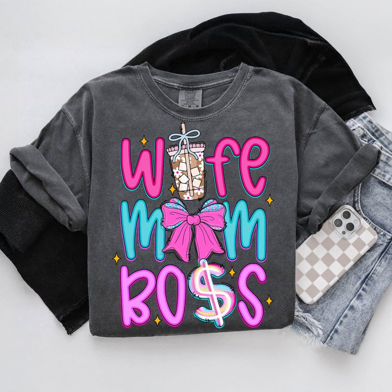 May include: Dark gray long-sleeve shirt with the words "Wife Mom Boss" in pink and blue, along with a graphic of a coffee drink and a pink bow. A phone with a checkered case and denim shorts are next to the shirt.