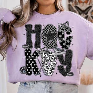 Peut inclure: Un sweat-shirt lilas avec le mot "HOCKEY" dans un m&eacute;lange de motifs, dont un imprim&eacute; l&eacute;opard, des pois et une &eacute;toile &agrave; carreaux. Le design comprend de l'&eacute;quipement de hockey comme des patins, un casque et des palets.