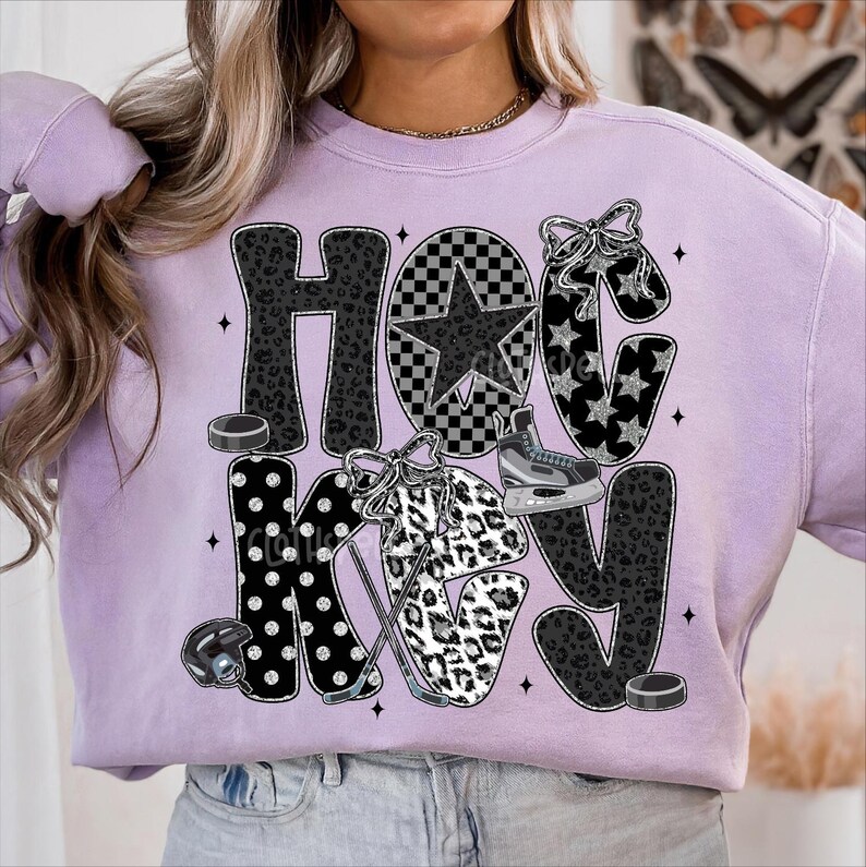 Peut inclure: Un sweat-shirt lilas avec le mot "HOCKEY" dans un m&eacute;lange de motifs, dont un imprim&eacute; l&eacute;opard, des pois et une &eacute;toile &agrave; carreaux. Le design comprend de l'&eacute;quipement de hockey comme des patins, un casque et des palets.