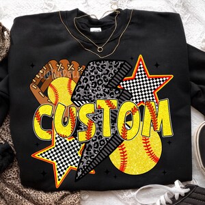 May include: Black sweatshirt with a softball theme. The design includes a softball, glove, and stars. The word "CUSTOM" is in yellow with a black outline. The lightning bolt has a leopard print design.