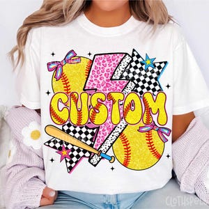 May include: White t-shirt with a colorful softball-themed design. The design includes yellow softballs, a baseball bat, and the word "CUSTOM" in yellow bubble letters. Accents include pink leopard print, checkered patterns, and bows.