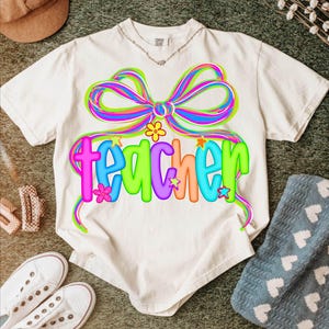 May include: White t-shirt with a colorful bow and the word "teacher" in a playful font. The design features bright colors like pink, blue, green, and orange, with floral and star accents. A casual, fun design.
