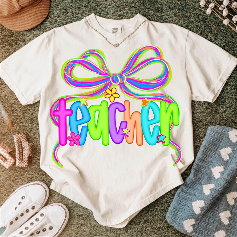 May include: White t-shirt with a colorful bow and the word "teacher" in a playful font. The design features bright colors like pink, blue, green, and orange, with floral and star accents. A casual, fun design.