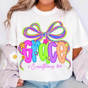 Puede incluir: Camiseta blanca con un dise&ntilde;o gr&aacute;fico colorido. La palabra "GRACE" se muestra en letras de burbujas vibrantes y multicolores, adornadas con estrellas y flores. Un gran lazo de arco&iacute;ris se encuentra encima de la palabra, con el texto "2 Corintios 12:9" debajo.