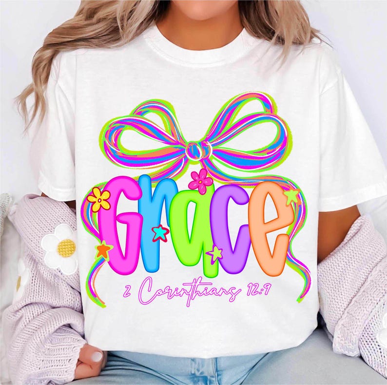 Puede incluir: Camiseta blanca con un dise&ntilde;o gr&aacute;fico colorido. La palabra "GRACE" se muestra en letras de burbujas vibrantes y multicolores, adornadas con estrellas y flores. Un gran lazo de arco&iacute;ris se encuentra encima de la palabra, con el texto "2 Corintios 12:9" debajo.