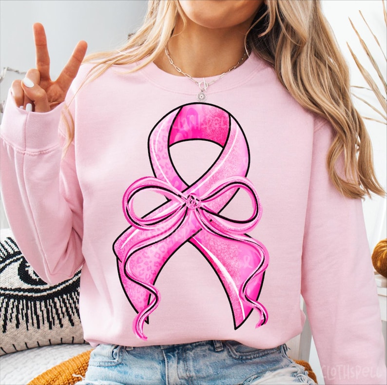 May include: A light pink sweatshirt featuring a large pink ribbon design with a black outline. The ribbon has a bow at the bottom and a leopard print pattern. The sweatshirt is a solid light pink color.