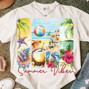 K&ouml;nnte beinhalten: Cremefarbenes T-Shirt mit einem farbenfrohen Strandmotiv. Das Design zeigt einen Strandball, ein Surfbrett, Palmen, Muscheln und den Text "Summer Vibes" in roter Schrift. Das T-Shirt hat einen Rundhalsausschnitt und kurze &Auml;rmel.