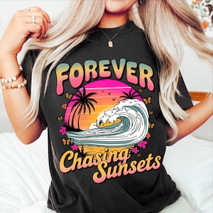 K&ouml;nnte beinhalten: Schwarzes T-Shirt mit einem farbenfrohen Grafikdesign. Das Design zeigt die W&ouml;rter "FOREVER" und "Chasing Sunsets" &uuml;ber und unter einer Sonnenuntergangsszene mit Palmen, Schmetterlingen und einer Welle.