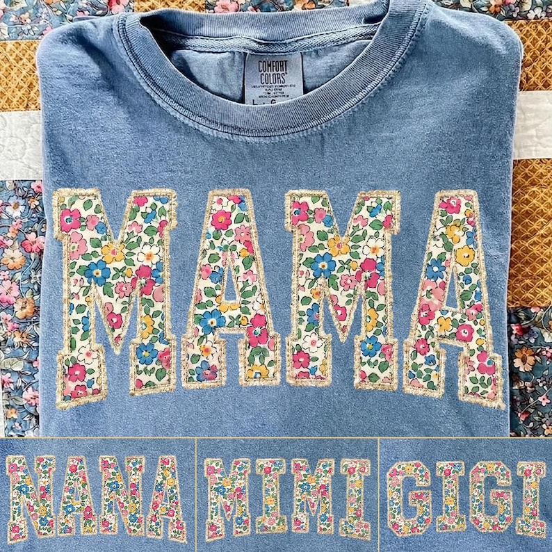 K&ouml;nnte beinhalten: Hellblaues Comfort Colors T-Shirt mit dem Wort "MAMA" in gro&szlig;en Buchstaben mit Blumenmuster. Die Buchstaben sind goldfarben umrandet. Darunter kleinere Textoptionen mit "NANA", "MIMI" und "GIGI" im gleichen Stil.