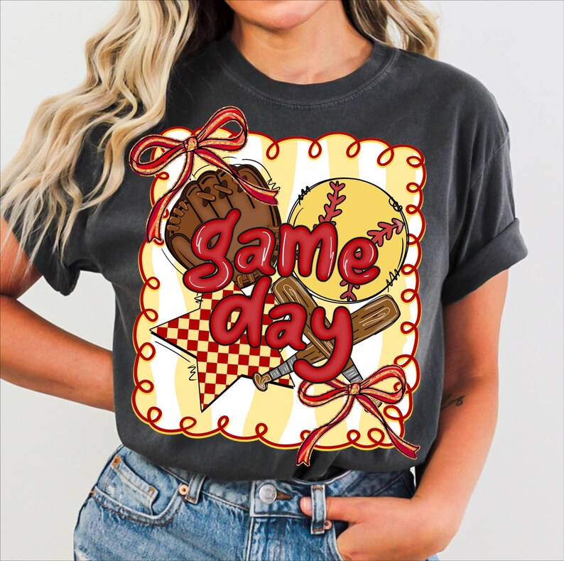 Puede incluir: Camiseta gris oscuro con un dise&ntilde;o tem&aacute;tico de softbol. El dise&ntilde;o incluye una pelota de softbol, un bate de b&eacute;isbol, un guante y una estrella con las palabras "Game Day" en rojo. El dise&ntilde;o est&aacute; enmarcado con un borde amarillo y lazos rojos.