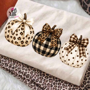May include: A white t-shirt featuring three pumpkin illustrations. The first is a leopard print pumpkin with a cream bow. The second is a black and white buffalo plaid pumpkin with a leopard print bow. The third is a white pumpkin with brown polka dots and a brown plaid bow.