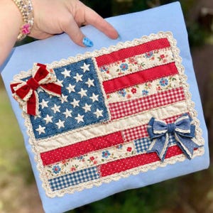 Puede incluir: Un coj&iacute;n hecho a mano con un dise&ntilde;o de patchwork de la bandera estadounidense. La bandera incluye un campo de mezclilla azul con estrellas blancas, rayas rojas y blancas y lazos decorativos. El coj&iacute;n est&aacute; adornado con encaje y sobre un fondo azul claro.