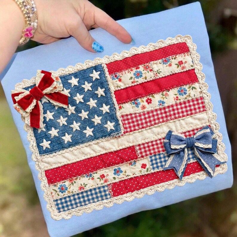 Puede incluir: Un coj&iacute;n hecho a mano con un dise&ntilde;o de patchwork de la bandera estadounidense. La bandera incluye un campo de mezclilla azul con estrellas blancas, rayas rojas y blancas y lazos decorativos. El coj&iacute;n est&aacute; adornado con encaje y sobre un fondo azul claro.