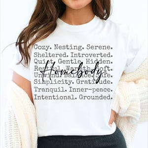 Peut inclure: T-shirt blanc avec du texte noir qui dit "Cozy. Nesting. Serene. Sheltered. Introverted. Quiet. Gentle. Hidden. Restful. Warmth. Soft. Unwind. Balance. Safe. Simplicity. Gratitude. Tranquil. Inner-peace. Intentional. Grounded." Le mot "Homebody" est &eacute;crit en plus grand et est centr&eacute; sur le t-shirt.