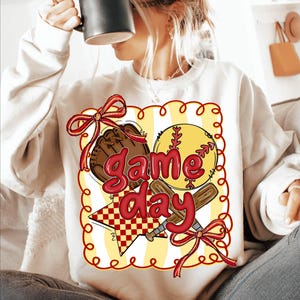 Puede incluir: Sudadera crema con un dise&ntilde;o de b&eacute;isbol. El gr&aacute;fico incluye una pelota de b&eacute;isbol, un guante y bates cruzados, con las palabras "Game Day" en rojo. El dise&ntilde;o est&aacute; enmarcado por un borde amarillo con detalles rojos.