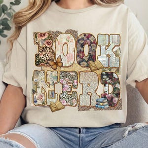 May include: Beige t-shirt with the words "BOOK NERD" in a decorative font. The letters are filled with floral, leopard print, and book page patterns. A butterfly and small books are also featured.