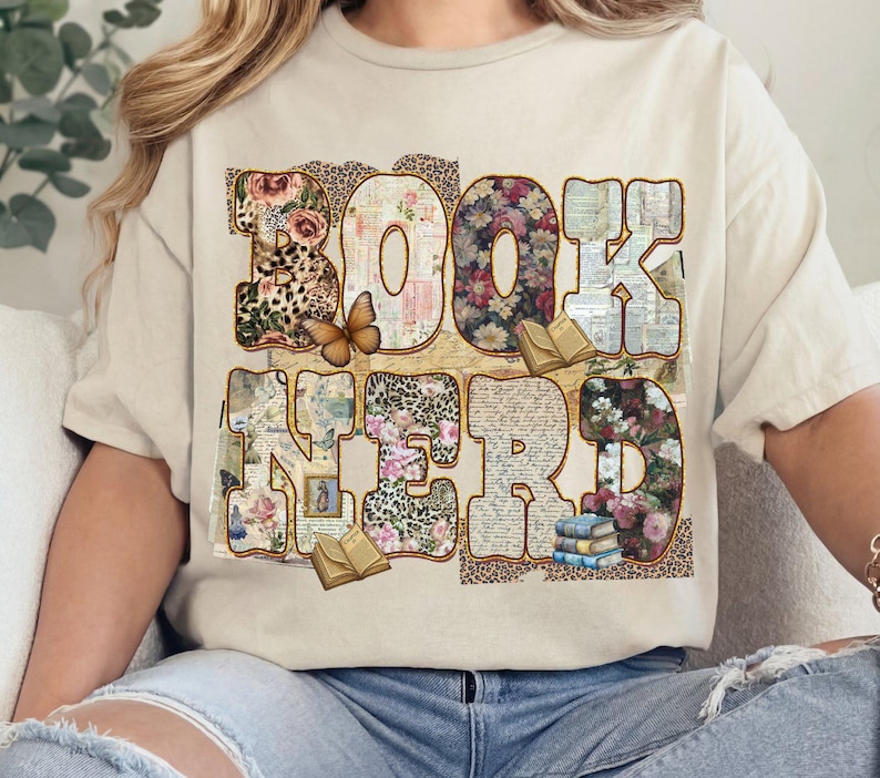 May include: Beige t-shirt with the words "BOOK NERD" in a decorative font. The letters are filled with floral, leopard print, and book page patterns. A butterfly and small books are also featured.
