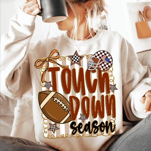 May include: Cream-colored sweatshirt with a football-themed graphic. The design includes a football, helmet, and the words "TOUCH DOWN season" in brown and black. The graphic is decorated with stars and a bow.