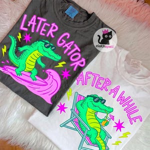 May include: Two t-shirts with alligator graphics. One is dark gray with the text "LATER GATOR" and a green alligator on a pink wave. The other is white with "AFTER A WHILE" and an alligator in a beach chair.