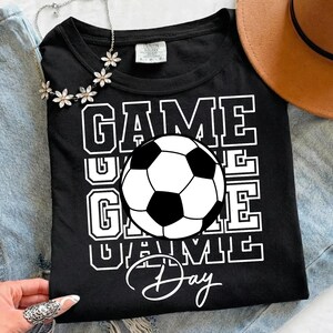 Może przedstawiać: Czarny t-shirt z białą grafiką piłki nożnej i napisem "GAME DAY" pogrubioną, blokową czcionką. Koszulka ma klasyczny okrągły dekolt, idealna dla fan&oacute;w sportu.