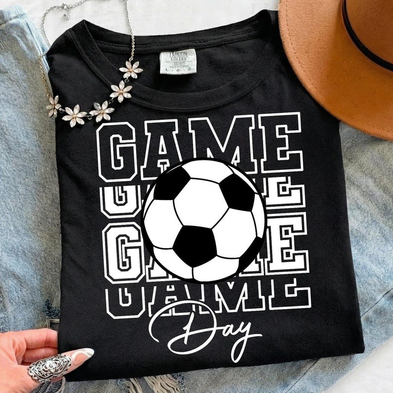 Może przedstawiać: Czarny t-shirt z białą grafiką piłki nożnej i napisem "GAME DAY" pogrubioną, blokową czcionką. Koszulka ma klasyczny okrągły dekolt, idealna dla fan&oacute;w sportu.