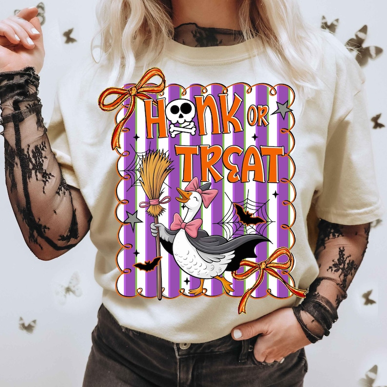 May include: Cream-colored t-shirt with a Halloween-themed graphic. The graphic features a goose wearing a cape and holding a broom, with the words "Honk or Treat" in orange and white. The design includes bats, spiderwebs, and a skull.