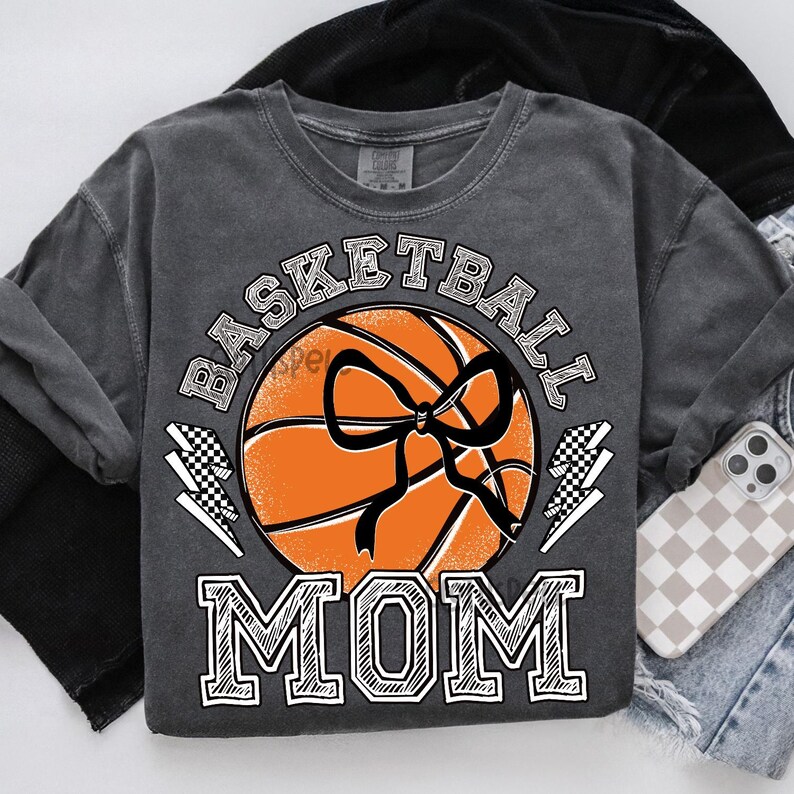 Pode incluir: Camisola cinza escuro com as palavras "BASKETBALL MOM" em branco. Uma bola de basquete laranja com um la&ccedil;o preto est&aacute; no centro. Raios quadriculados ladeiam a bola. Um telefone em uma capa quadriculada &eacute; vis&iacute;vel.