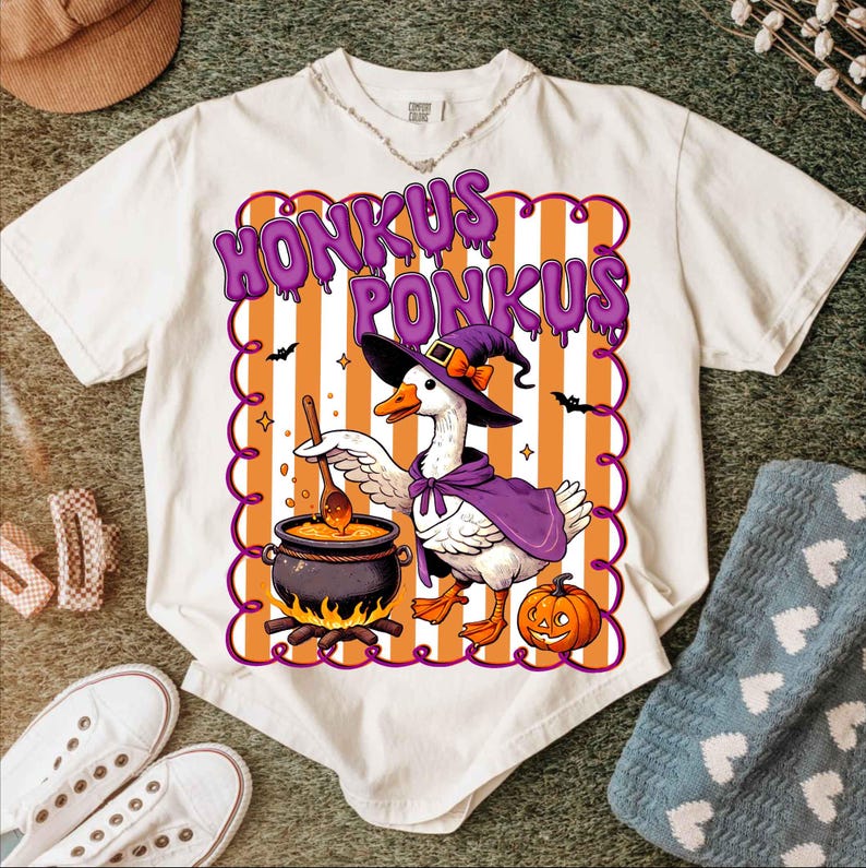 May include: White t-shirt with a Halloween-themed graphic. The design features a goose in a witch's hat and cape, stirring a cauldron. The text "HONKUS PONKUS" is displayed above the goose in purple letters.