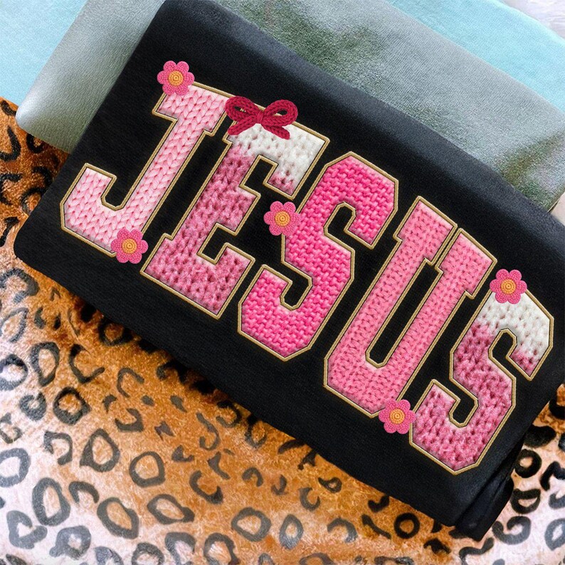 May include: Black fabric with the word "JESUS" embroidered in pink and white, outlined in gold. The letters have a knitted texture and are decorated with small pink flowers and a red bow.