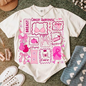 May include: Cream-colored t-shirt with a pink graphic design related to breast cancer awareness. The design includes the words "Breast", "Fight", "Hope", and "Cure", along with pink ribbons, a bra, a pumpkin, and other related images.