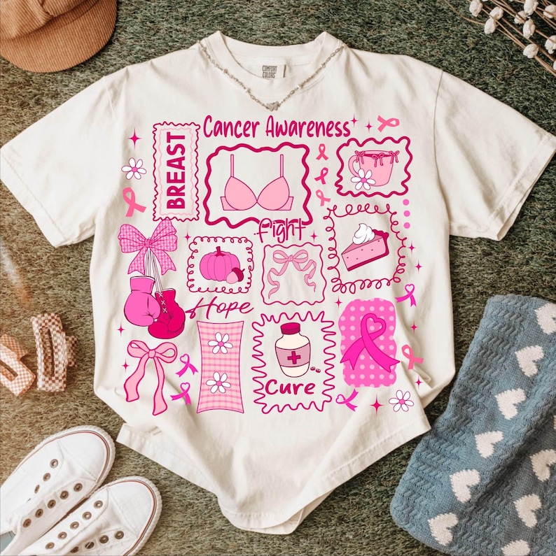 May include: Cream-colored t-shirt with a pink graphic design related to breast cancer awareness. The design includes the words "Breast", "Fight", "Hope", and "Cure", along with pink ribbons, a bra, a pumpkin, and other related images.