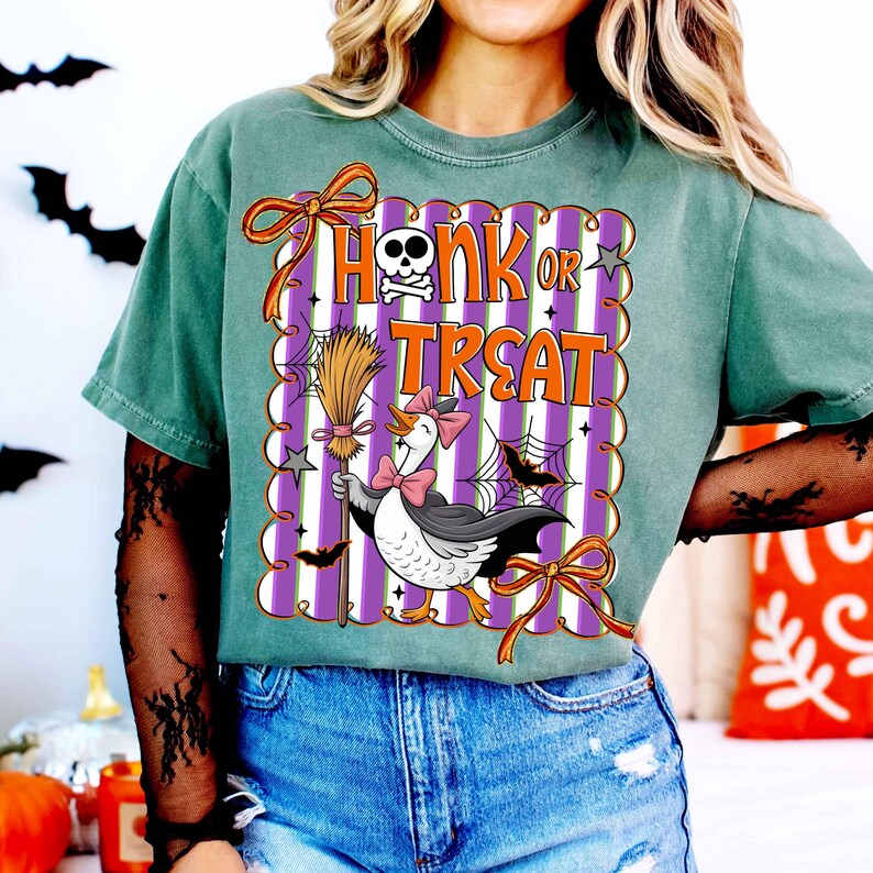 May include: Green t-shirt with a Halloween-themed graphic. The graphic features a goose wearing a bow tie and holding a broom, with the words "Honk or Treat" in orange. The design is set against a purple and white striped background.