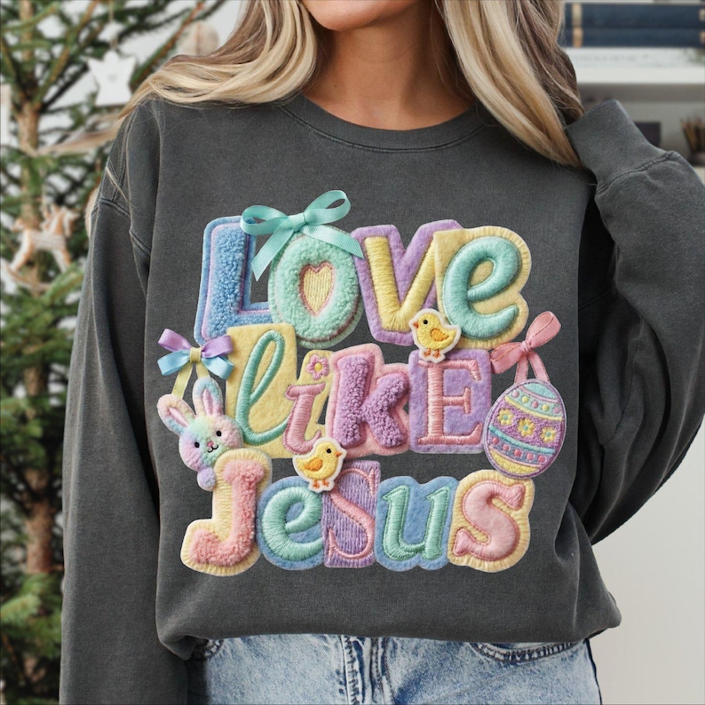 Pu&ograve; includere: Felpa grigio scuro con la scritta "Love Like Jesus" in lettere colorate e strutturate. Il design include un coniglietto, pulcini, un uovo di Pasqua e fiocchi, il tutto in colori pastello.