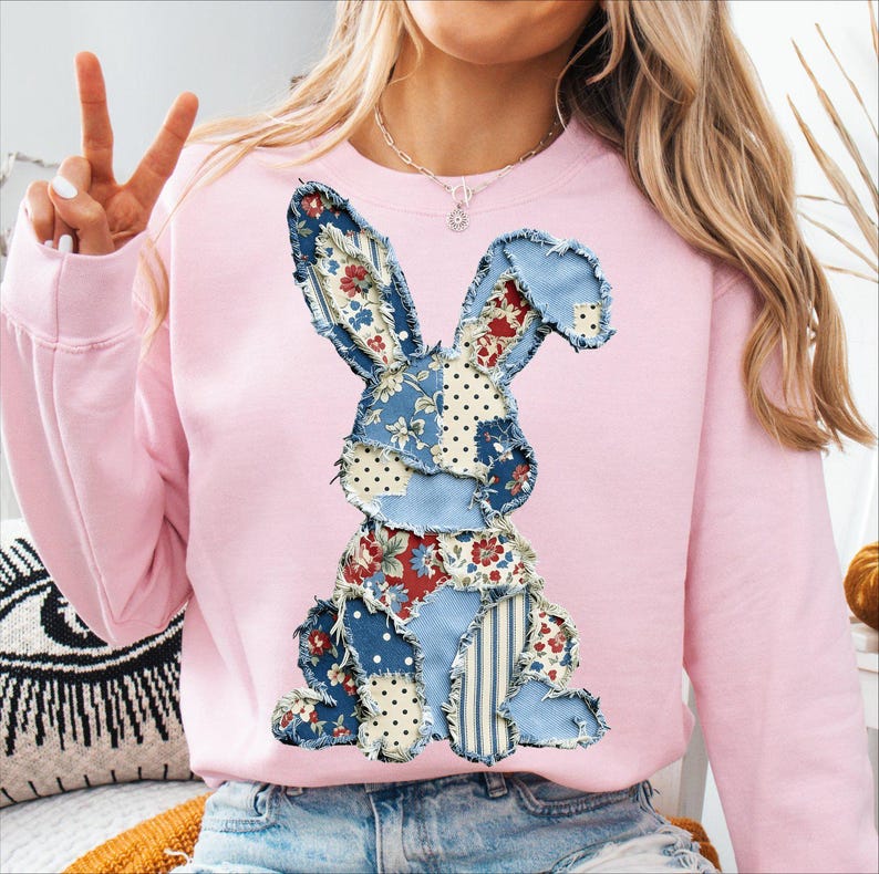 May include: A light pink sweatshirt featuring a patchwork bunny design. The bunny is made of various blue fabrics with floral, polka dot, and striped patterns. The sweatshirt has long sleeves and a crew neck.