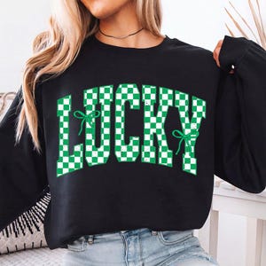 May include: Black sweatshirt with the word "LUCKY" in large, green and white checkered letters. The word "LUCKY" has green bow accents. The sweatshirt is a long-sleeved crew neck.