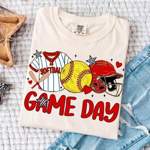 May include: Cream-coloured t-shirt with a softball design. The design includes a softball, a red and white softball jersey, a red helmet, and the words "GAME DAY" in red. Accents include hearts and stars.