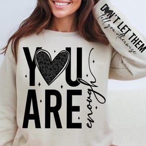 Peut inclure: Un sweat-shirt beige avec le texte "YOU ARE enough" en lettres noires. Un c&oelig;ur noir avec un motif l&eacute;opard est au centre du texte. Le texte "DON'T LET THEM tell you otherwise" est sur la manche.