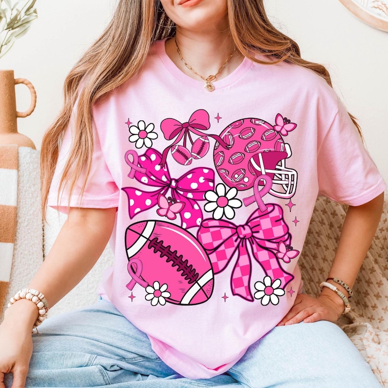 May include: Pink t-shirt featuring a football theme with pink bows, footballs, a helmet, and daisies. The design includes a pink ribbon, butterflies, and stars. The shirt is a light pink color.