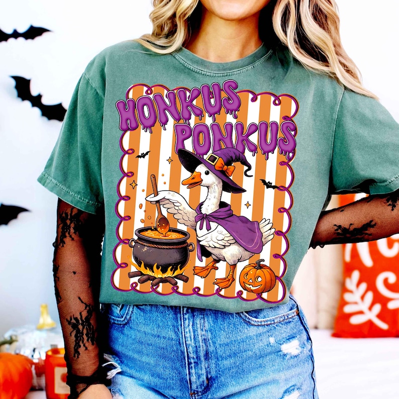 May include: Green t-shirt with a Halloween-themed graphic. The graphic features a goose wearing a witch hat and cape, stirring a cauldron. The text "HONKUS PONKUS" is above the goose. A pumpkin and bats are also included in the design.