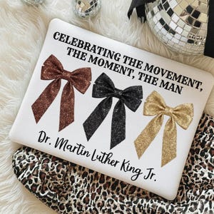 May include: White pillow with three glittery bow ties in brown, black, and gold. Text reads "CELEBRATING THE MOVEMENT, THE MOMENT, THE MAN" and "Dr. Martin Luther King Jr." in black font. The pillow is on a leopard print fabric.