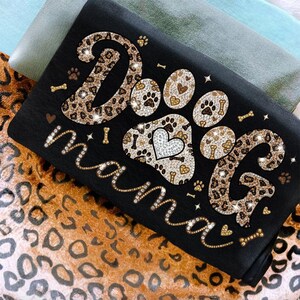 May include: Black t-shirt with the words "Dog Mama" in a sparkling, cursive font. The word "Dog" is in a leopard print design, and the paw prints have a heart and bone pattern. The shirt is folded on a leopard print fabric.